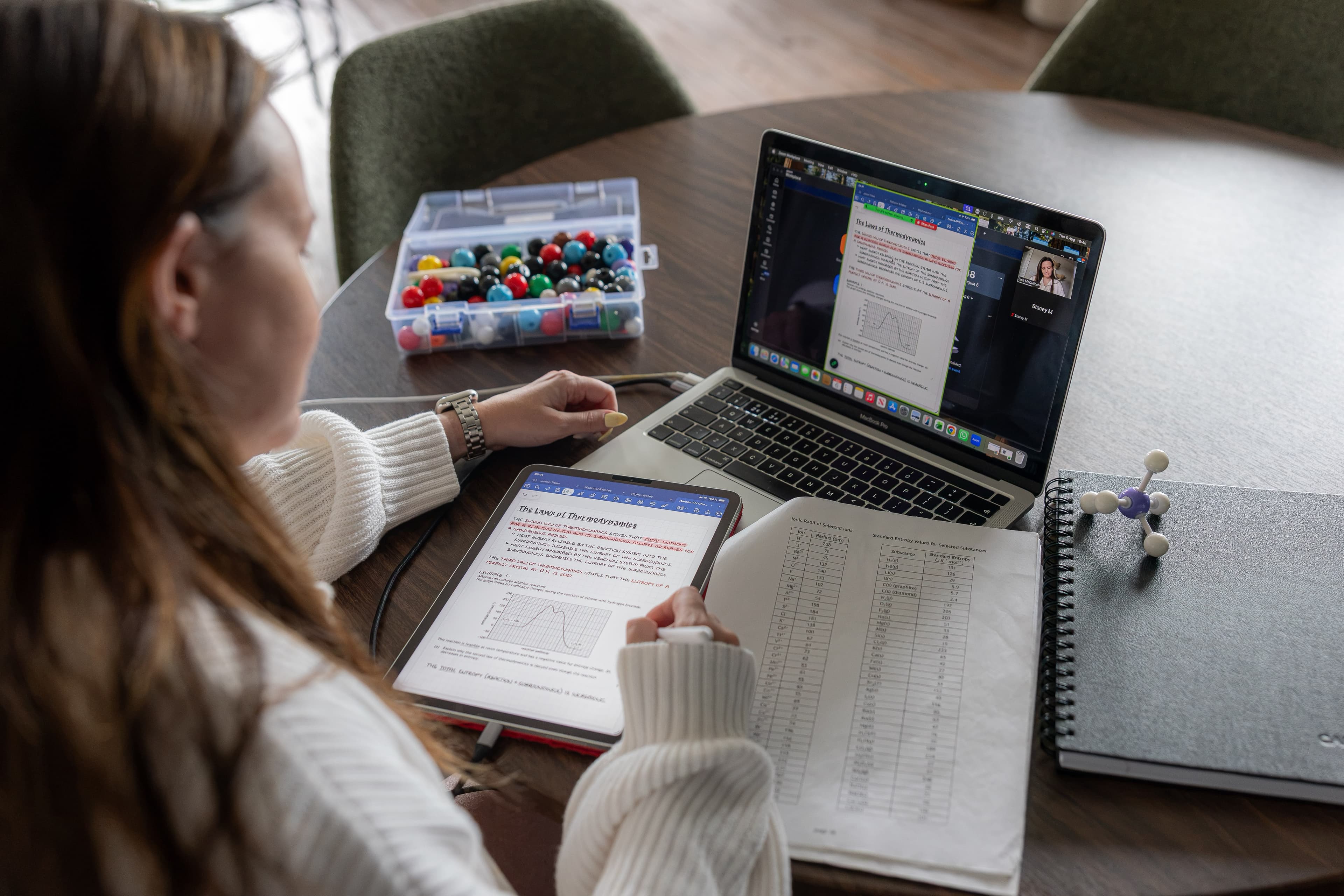 A pupil following a live online Chemistry lesson on a laptop, annotating notes on a tablet beside past-paper data sheets
